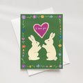 A greeting card with a green background, featuring two cream-colored rabbits facing each other beneath a pink heart that reads 'I love you.' The card is decorated with a border of small flowers and green vines.