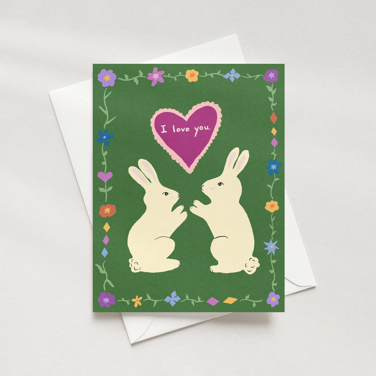 A greeting card with a green background, featuring two cream-colored rabbits facing each other beneath a pink heart that reads 'I love you.' The card is decorated with a border of small flowers and green vines.