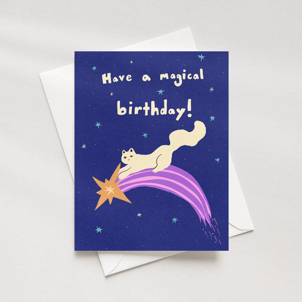 A birthday card with a dark blue night-sky design, featuring a cream-colored cat riding a purple comet with an orange star tail. The text reads 'Have a magical birthday!'.
