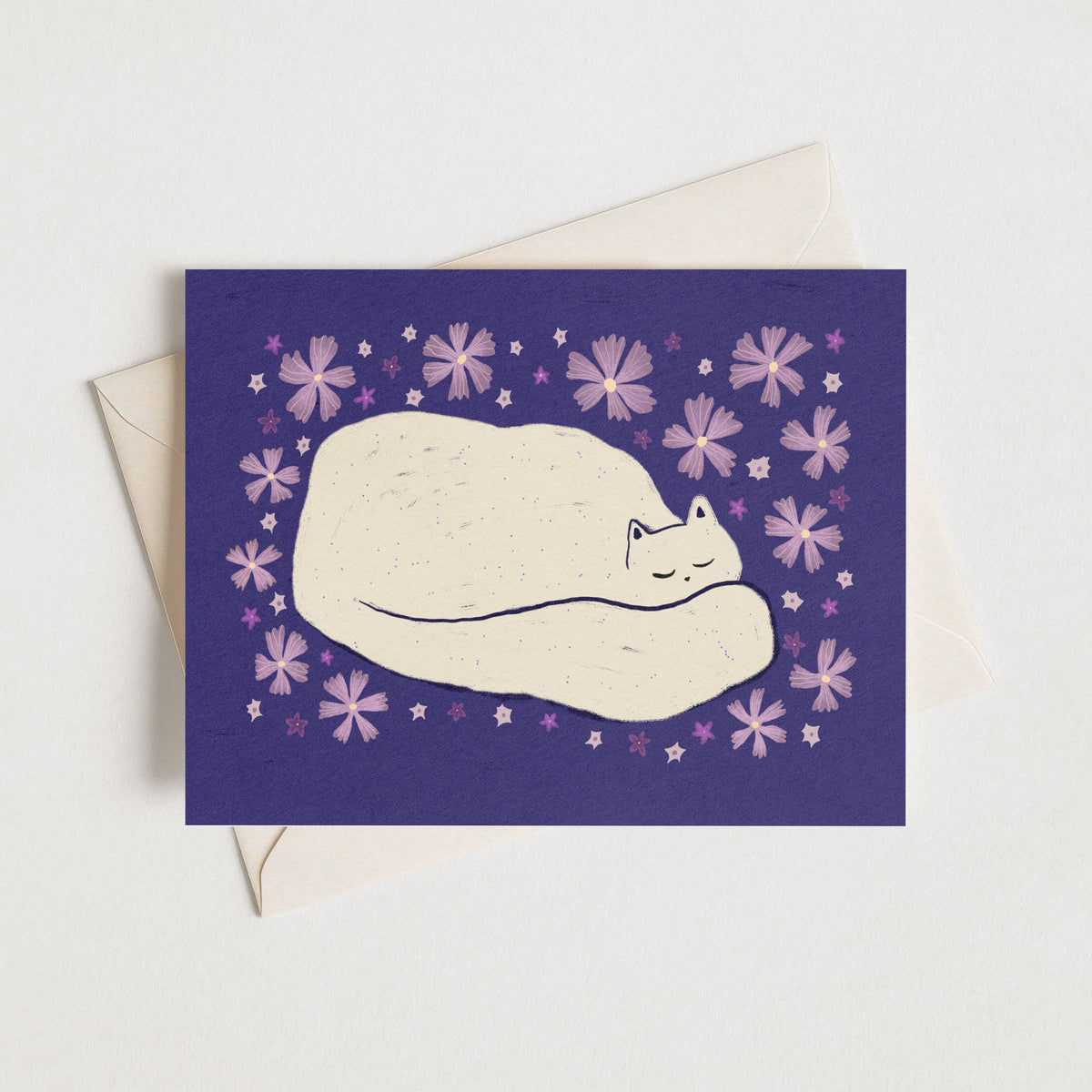 A square greeting card with a purple background, featuring a cream-colored sleeping cat surrounded by small purple flowers, shown with a cream envelope on a light neutral background.
