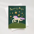 A greeting card with a whimsical unicorn illustration and the words 'Sending Magic' in gold, shown with a white envelope on a light neutral background.