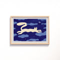 An art print featuring a whimsical illustration of a cream-colored cat stretching across a deep blue night sky with clouds, stars, and a crescent moon, displayed on a clean white wall.