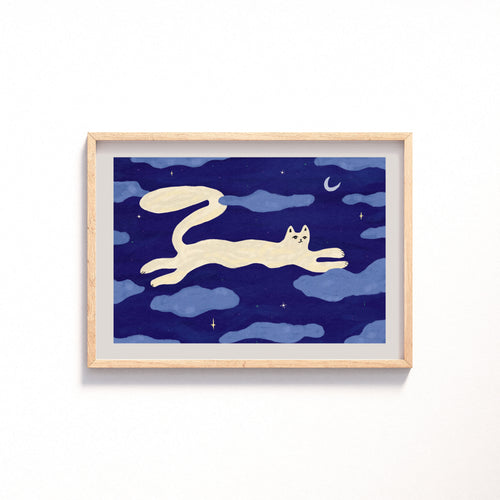 An art print featuring a whimsical illustration of a cream-colored cat stretching across a deep blue night sky with clouds, stars, and a crescent moon, displayed on a clean white wall.